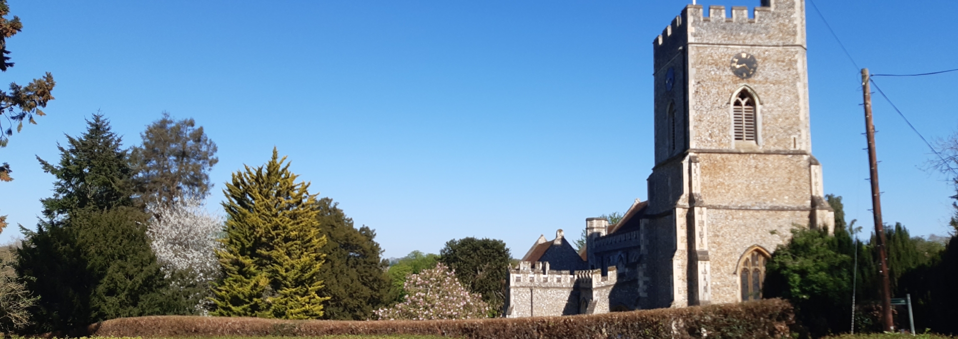 Stone church with a clock tower, surrounded by trees and hedges, under a clear blue sky.